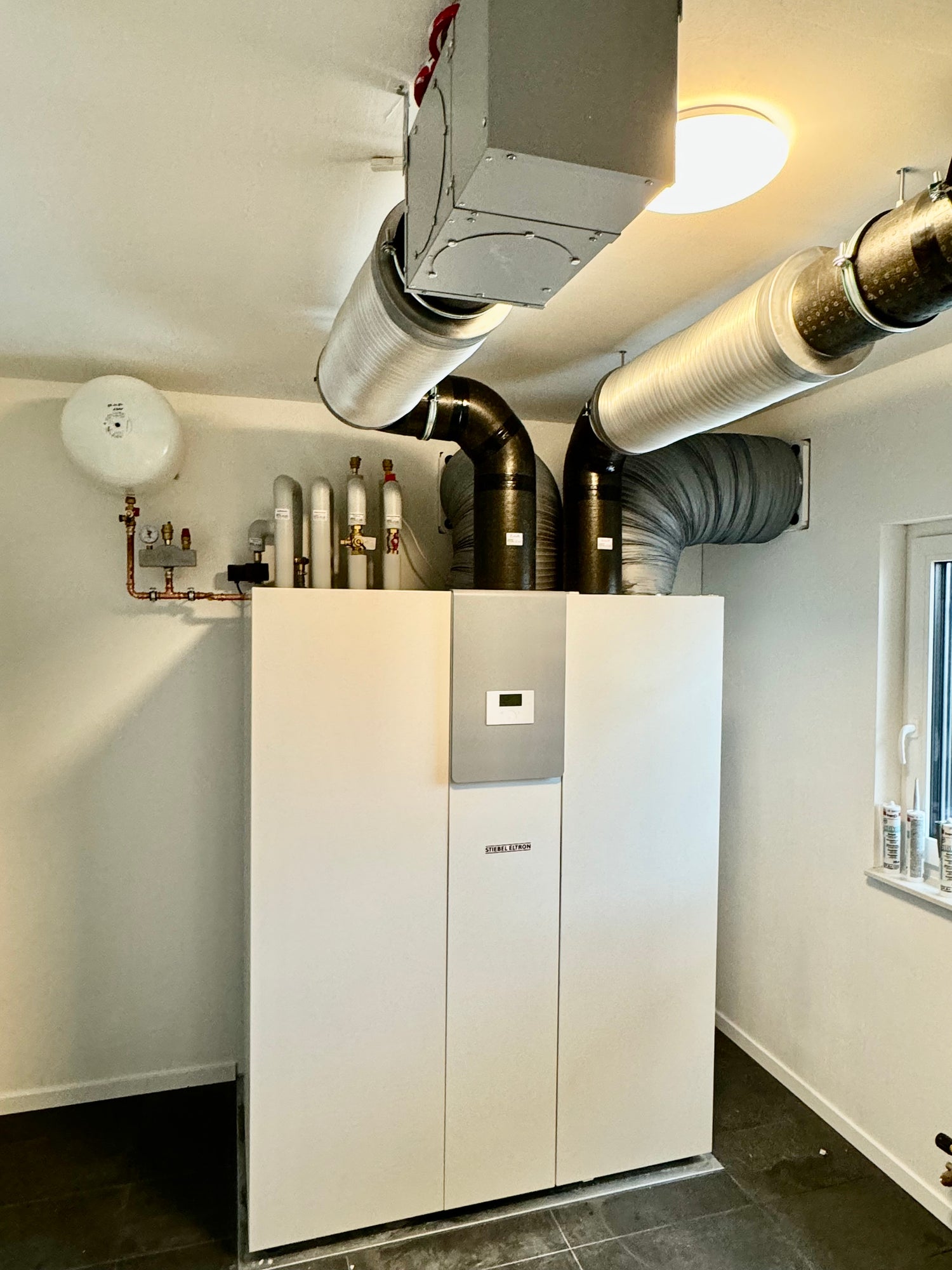 Im Bild ist ein Heizungsraum eines Neubaus zu sehen. Es ist eine Wärmepumpe mit integrierter Lüftung von Stiebel Eltron verbaut worden. Im oberen Bereich sind sowohl Kupferleitungen für die Heizung und Warmwasser zu sehen, als auch die Schalldämpfer und Verteilerboxen der Lüftungsanlage