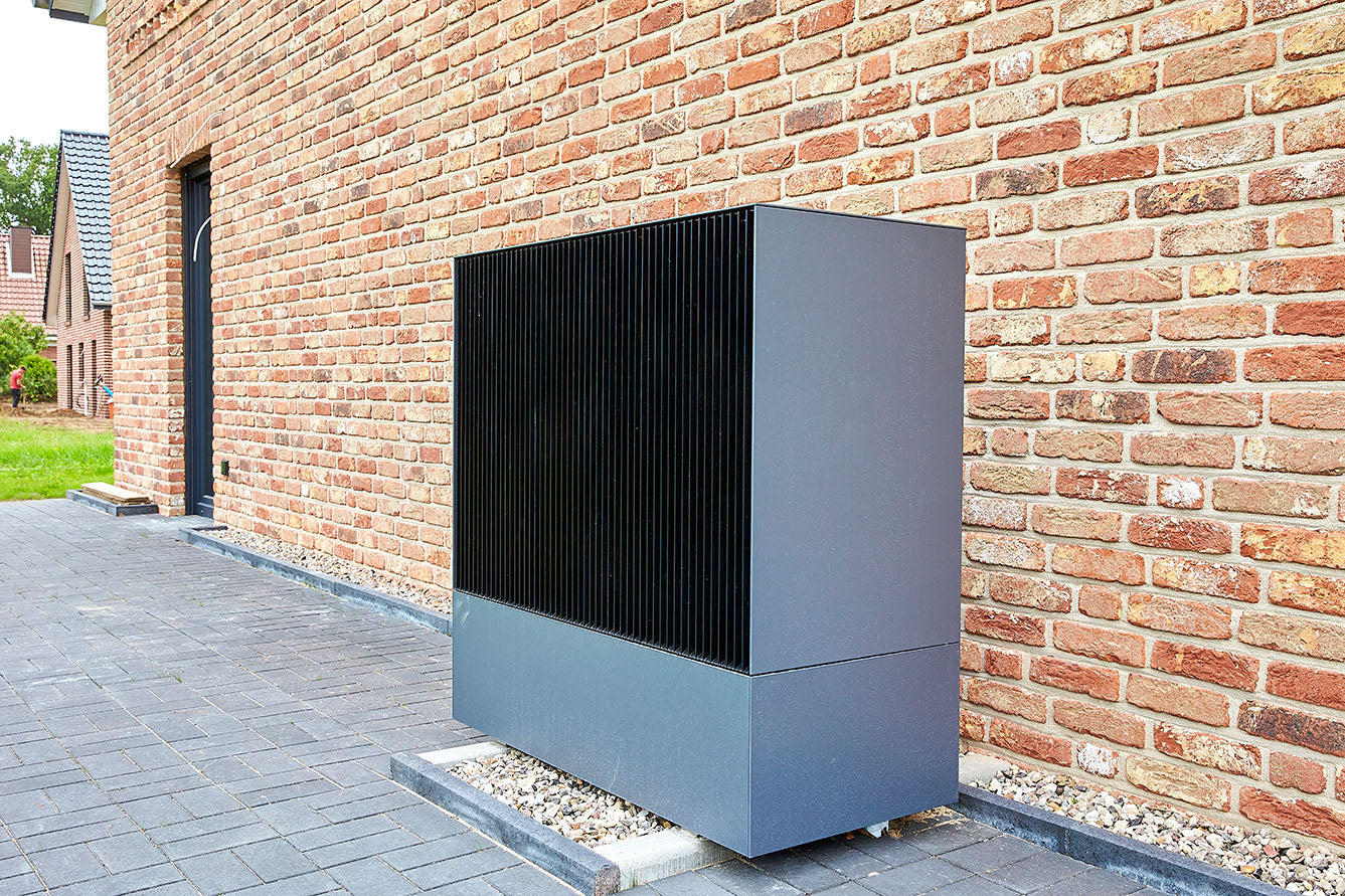 Auf diesem Bild ist eine Viessmann Wärmepumpe vor einem verklinkerten Gebäude zu sehen. Unter der Wärmepumpe ist ein Kiesbett, sodass Kondenswasser abtropfen kann. Der Hof-Vorbereich ist mit grauem Pflaster gepflastert.